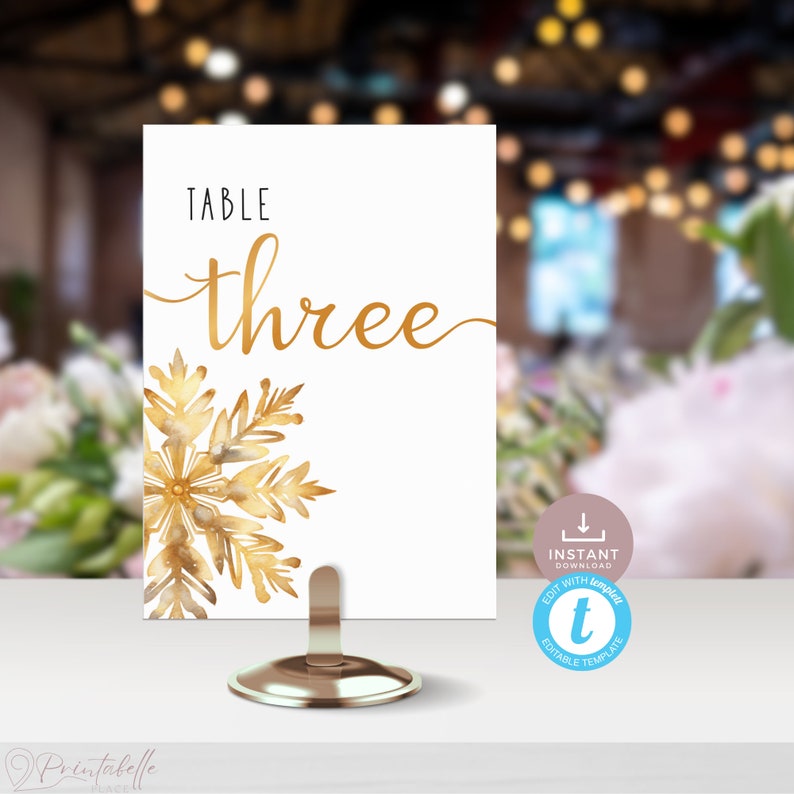 Table Number Card in a short gold stand with blurred dinner party scene in background. Card features a white background with a single large, detailed, intricate snowflake in gold tones. It sits in bottom left with about three quarters of it visible.