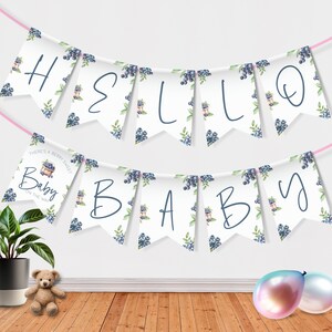 Op de afbeelding: Een witte banner met blauwe en groene bosbessenontwerpen en de woorden "Hello Baby" in cursief. De banner is vastgemaakt met roze touw en hangt aan een witte muur.