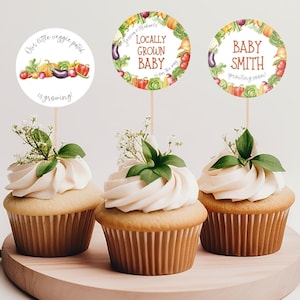 May include: Three cupcakes decorated with white frosting and green leaves. Each cupcake has a round, colorful sticker with a vegetable theme. The stickers say "Our little veggie patch is growing!" "Jessien & Michaels Locally Grown Baby is on the way" and "Baby Smith Sprouting Soon!"