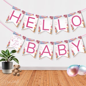 Op de afbeelding: Een roze en witte banner met de woorden "Hello Baby" en "Carmen's Ciclita Linda is soon due" in een bloemig ontwerp. De banner hangt aan een witte muur met een teddybeer en ballonnen op de voorgrond.