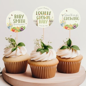 May include: Three cupcakes decorated with green leaves and white frosting. Each cupcake has a round, white paper topper with a garden theme. The toppers say "Baby Smith Sprouting Soon!", "Locally Grown Baby is on the way", and "Something's Growing".