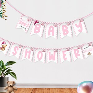 Op de afbeelding: Roze en witte slinger voor een babyshower. De slinger spelt "BABY SHOWER" in grote roze letters. Extra banners lezen "Merry little baby on the way!" en tonen een teddybeer met een kerstmuts.