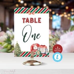 May include: Red and green striped table number card with the text "TABLE One" in white and red. The card features a Christmas tree and two cartoon sweaters with a snowman and reindeer design. The card is on a silver stand.