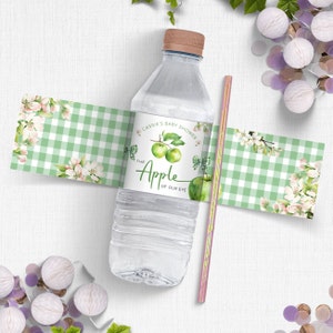 Op de afbeelding: Een groen en wit geruit waterfleslabel met een aquarel-illustratie van appels en bloesems. Het label luidt "Cassie's Baby Shower The Apple of Our Eye".