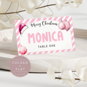 May include: Pink and white striped Christmas place card with a Santa hat and ornaments. The card says "Merry Christmas" and "Monica, Table One".