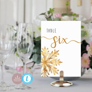 Final image of a table number in a short gold holder. It sits on a set table in front of a centerpiece of a vase of flowers. A place setting with wine glass, cutlery and crockery sits just to the left.