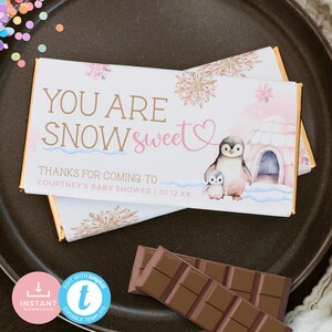 May include: A white chocolate bar wrapper with a pink and gold snowflake design. The wrapper features a penguin family and the text "YOU ARE SNOW sweet" and "THANKS FOR COMING TO COURTNEY'S BABY SHOWER | 01.12.XX".