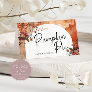 May include: A printable food label with a watercolor background in shades of orange and brown. The label features a floral design with a white circle in the center. The text "Pumpkin Pie" is written in black cursive script with the words "Warm & Delicious" below it.