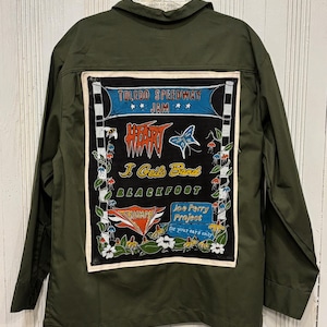 May include: Olive green jacket with a large graphic on the back featuring the text "Toledo Speedway Jam" and band names like "Heart" and "J. Geils Band." The graphic has a black background with white and colorful accents.