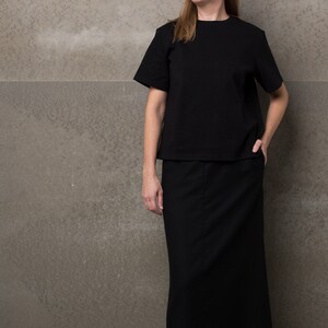 May include: A woman wearing a black top and a black maxi skirt. She is standing in front of a gray wall.