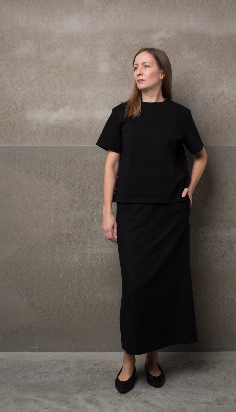 May include: A woman wearing a black top and a black maxi skirt. She is standing in front of a gray wall.