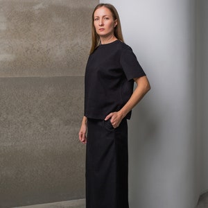 Peut inclure: Une femme portant un haut noir à manches courtes et une longue jupe noire. Elle se tient devant un mur gris et un mur blanc.