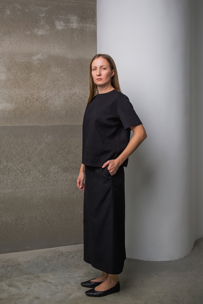May include: A woman wearing a black, short-sleeved top and a long black skirt. She is standing in front of a gray wall and a white wall.