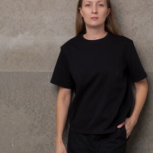 May include: A woman wearing a black t-shirt and a black skirt. She is standing in front of a gray wall.