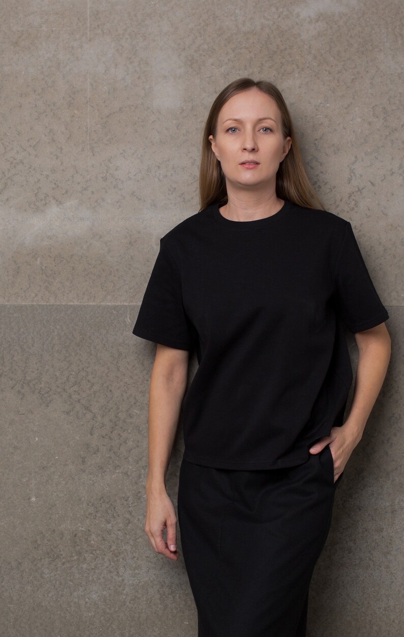 May include: A woman wearing a black t-shirt and a black skirt. She is standing in front of a gray wall.