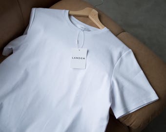 May include: A white t-shirt with a white tag that says "LANDEM" on a brown leather couch.