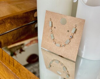 May include: A delicate necklace featuring rectangular, pale green gemstones and iridescent beads strung on a gold-colored chain. The necklace is displayed on a brown card, reflecting in a mirror. The design is simple and elegant.