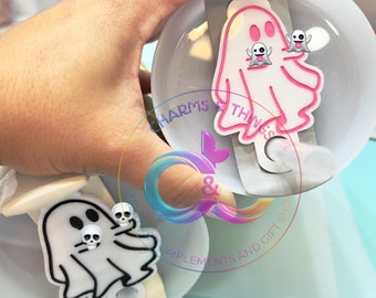 May include: Two white ghost-shaped bottle stoppers with black eyes and mouths. One ghost has a pink outline and the other has a black outline. The stoppers are on a white background.
