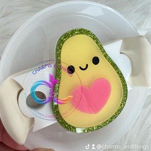 May include: A green glitter avocado charm with a pink heart and a smiling face. The charm is attached to a white plastic bottle stopper.