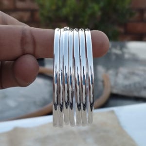 Peut inclure: Un ensemble de sept bracelets brillants en argent. La surface lisse et polie du métal reflète la lumière. Les bracelets sont tenus par les doigts d'une personne. Les bijoux sont de Jewelry By Priyanshi.