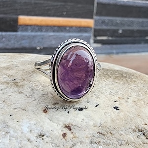 May include: A silver ring with a large oval amethyst gemstone. The ring has a decorative border around the stone.