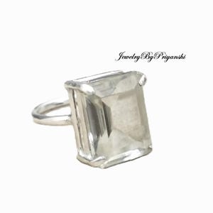 Op de afbeelding: Een zilveren ring met een grote, rechthoekige, heldere edelsteen. De ring heeft een eenvoudige band en een zetting die de edelsteen stevig vasthoudt. De tekst "Jewelry By Priyanshi" staat in de rechterbovenhoek.
