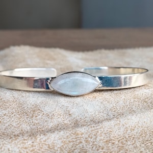May include: A silver bracelet with a white moonstone set in the center. The bracelet is open and has a smooth, polished finish.