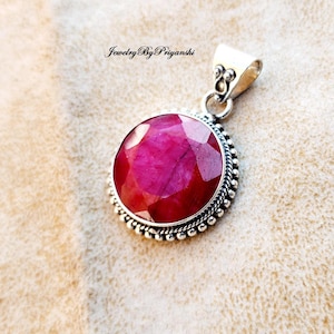May include: A round, faceted ruby pendant with a silver-colored setting. The pendant has a bail for a chain and a beaded border. The text "Jewelry By Priyanshi" is in the upper left corner.