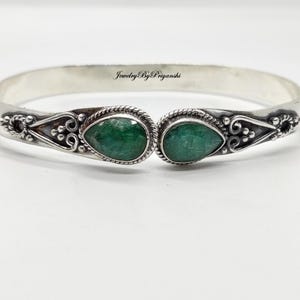 May include: A silver bangle bracelet with two emerald green stones set in the center. The bracelet has a delicate design with small swirls and beads.