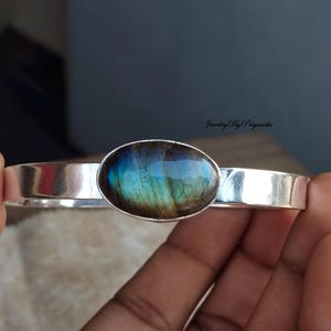 May include: A silver bangle bracelet with a large oval-shaped labradorite gemstone set in the center. The gemstone has a blue, green, and brown iridescent sheen.