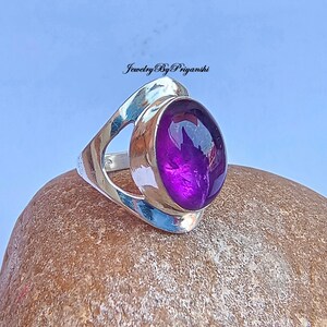 May include: A silver ring with an open design and a large, oval-shaped amethyst gemstone. The ring is set on a brown, textured surface. Jewelry By Priyanshi is written in the top center of the image.