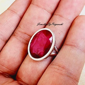 May include: A silver ring with a large, oval-shaped, red ruby gemstone. The ring is set in a simple, silver band.