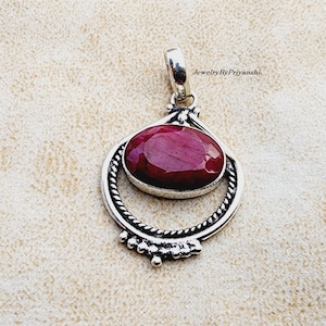 May include: A silver pendant with a deep red oval ruby gemstone set in a silver circle with a twisted rope design. The pendant has a silver bail for attaching to a chain.