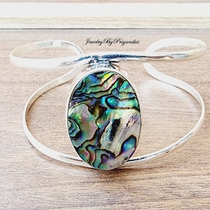 Puede incluir: Pulsera de plata con una incrustación ovalada de concha de abulón. La concha tiene colores iridiscentes de verde, azul y morado.