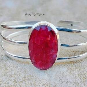 May include: A silver bangle bracelet with three bands and a large oval-shaped red gemstone set in the centre.