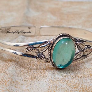 May include: Silver bangle bracelet featuring a light blue oval gemstone and leaf accents. The bracelet is engraved with the text "Jewelry By Priyanshi".