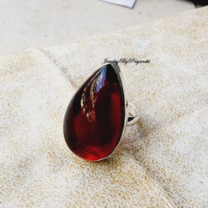 May include: A teardrop-shaped red gemstone ring set in silver. The ring is on a white fabric background.