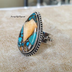 Peut inclure: Une bague en argent avec une pierre en turquoise et en corail fossilisé en forme de larme. La pierre est un mélange de couleurs bleues, vertes, blanches et orange. La bague a un bord perlé et un délicat motif floral sur la bande.