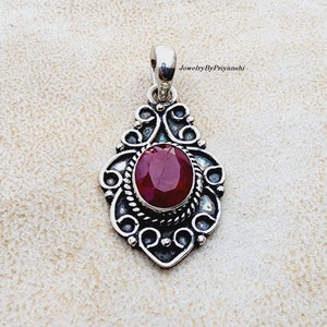 Puede incluir: Un colgante de plata con un diseño ornamentado que presenta una piedra preciosa de rubí rojo. El colgante tiene la forma de una lágrima y tiene un aro para sujetarlo a una cadena.