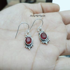 Könnte beinhalten: Ein Paar silberne Ohrhänger mit roten Edelstein-Akzenten. Die Ohrringe haben ein zartes Design mit kleinen Perlen und einem Hakenverschluss.