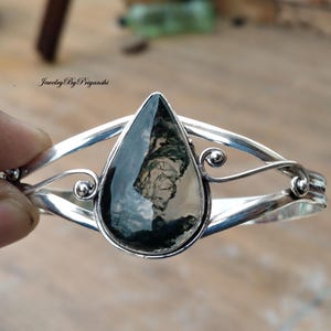 Puede incluir: Una pulsera de plata con una piedra de ágata verde musgo en forma de lágrima. La pulsera tiene un diseño delicado y adornado.