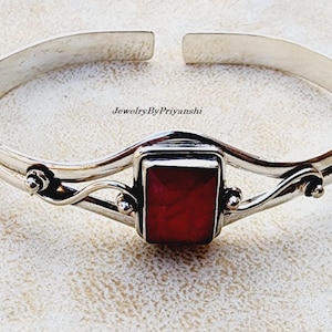 May include: A silver bangle bracelet with a red gemstone set in a silver frame. The bracelet has a delicate, twisted design and is accented with small silver beads.