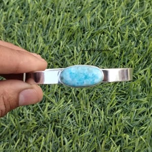 Puede incluir: Un brazalete de plata con una piedra ovalada de color azul claro engastada en el centro.