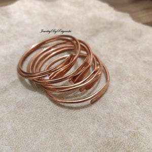 Puede incluir: Un conjunto de seis brazaletes de cobre apilados uno encima del otro. Los brazaletes tienen todos el mismo ancho y un acabado ligeramente martillado.