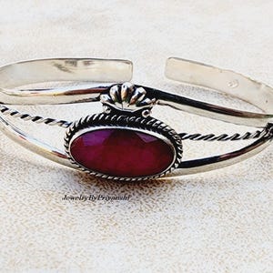 Peut inclure: Un bracelet jonc en argent avec une pierre précieuse rouge sertie dans un ovale en argent. Le bracelet est orné d'un motif de corde torsadée et d'un petit accent en forme de couronne.