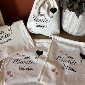 Può includere: Quattro sacchetti in cotone color crema con coulisse, caratterizzati dalla scritta nera Team Mariée seguita dai nomi Gwendoline, Emelyne, Valentin e Serena. Ogni sacchetto è decorato con un palloncino nero a forma di cuore e cuoricini rosa.