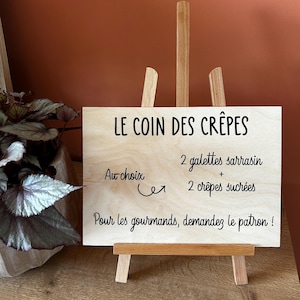 Peut inclure: Un panneau rectangulaire en bois clair posé sur un petit chevalet en bois. Le panneau comporte un texte imprimé en noir : LE COIN DES CRÊPES, 2 galettes sarrasin + 2 crêpes sucrées, Au choix, et Pour les gourmands, demandez le patron !