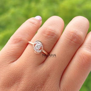 May include: A gold ring with a central diamond surrounded by smaller diamonds in an oval setting. The ring is on a person's finger, with a blurred green background. The ring has the word "NOAHA" on the side.