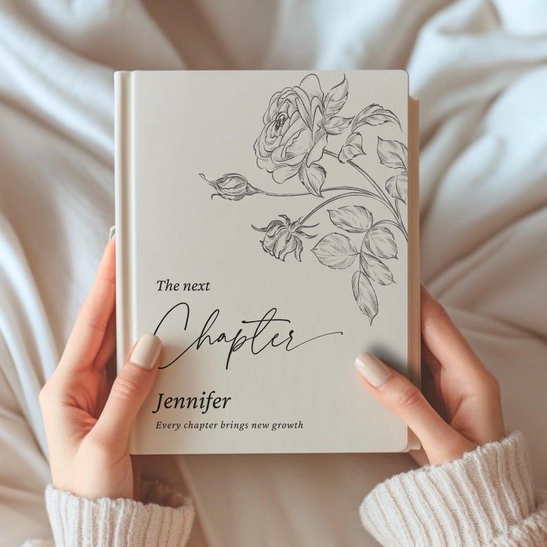 May include: A cream-colored book with a floral rose design and the words "The next Chapter" and "Jennifer" in elegant script. The book also includes the phrase "Every chapter brings new growth".