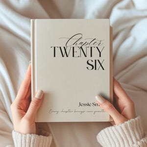 Peut inclure: Un livre à couverture rigide de couleur crème avec du texte noir sur la couverture. Le texte indique Chapter TWENTY SIX, Jessie Sm, et Every chapter brings new growth. Le livre est tenu droit, présentant un design épuré avec des polices serif.